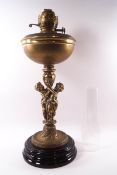 A Victorian brass oil lamp supported by two cast cherubs on black earthenware plinth,