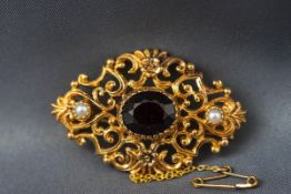 A yellow gold brooch set with a central oval garnet and two seed pearls. Ornate scroll design.