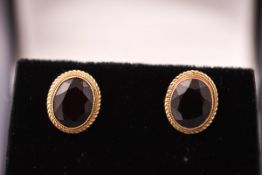 A yellow metal pair of stud earrings each set with an oval cut garnet. Post and butterfly fitting.