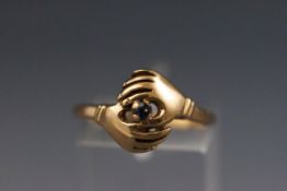 A yellow metal ring stylized as two hands set with a single sapphire.
