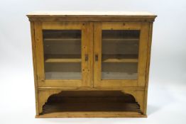 A pine wall hanging cabinet with glazed doors enclosing shelves above a rack, 104.