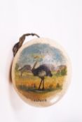An ostrich egg painted with a scene of a bird and with fabric hanging loop