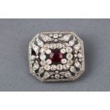 A white metal abstract brooch centrally set with an oval faceted ruby and surrounded by diamonds.