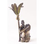 A gold painted bronze figure seated on a boulder holding a sword and shield, beneath a palm tree,