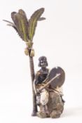 A gold painted bronze figure seated on a boulder holding a sword and shield, beneath a palm tree,