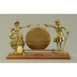 A brass dinner gong, suspended between two figures and raised on an oak base. Length 36 cm.