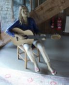 A life-size papier-mache figure of a girl seated upon a chair playing guitar,