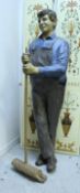 A life-size papier-mache figure of a man in dungarees leaning upon a broom,