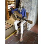 A life-size papier-mache figure of a girl seated upon a chair playing guitar,