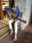 A life-size papier-mache figure of a girl seated upon a chair playing guitar,
