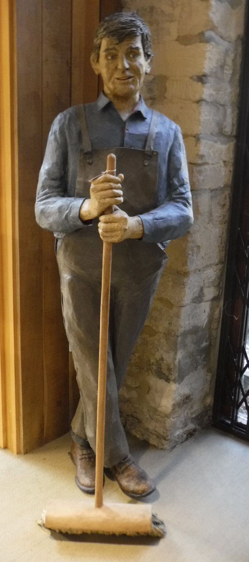 A life-size papier-mache figure of a man in dungarees leaning upon a broom,