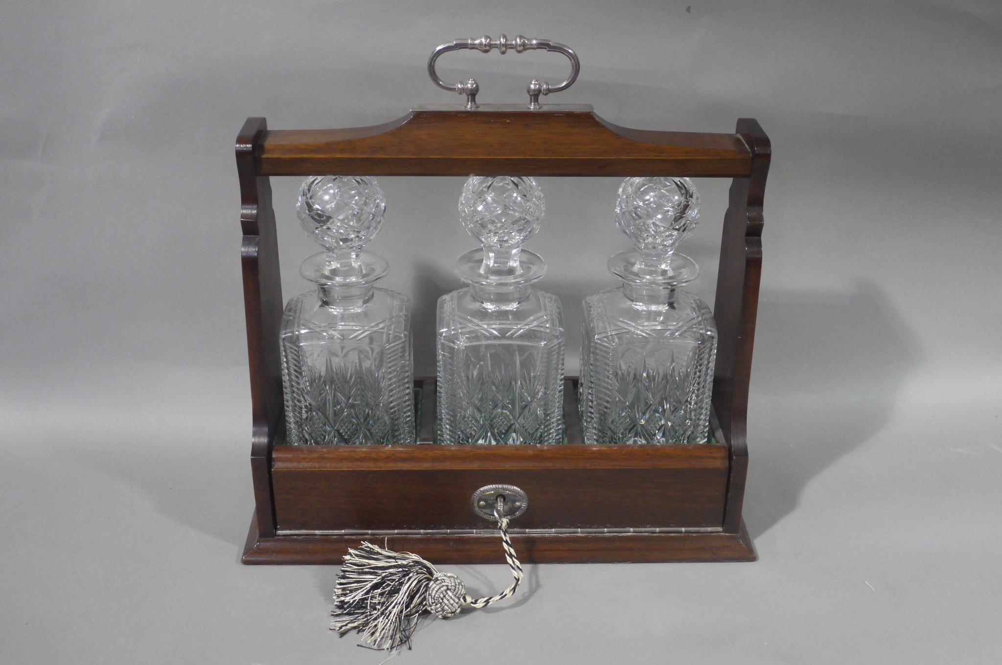 A reproduction mahogany finish three bottle Tantalus with silver plated loop carrying handle