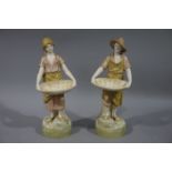 A pair of Royal Dux figures of a young boy and girl both holding a woven basket on rustic circular