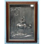 A silvered cast metal relief plaque of a mounted camel under a palm tree, glazed and framed,