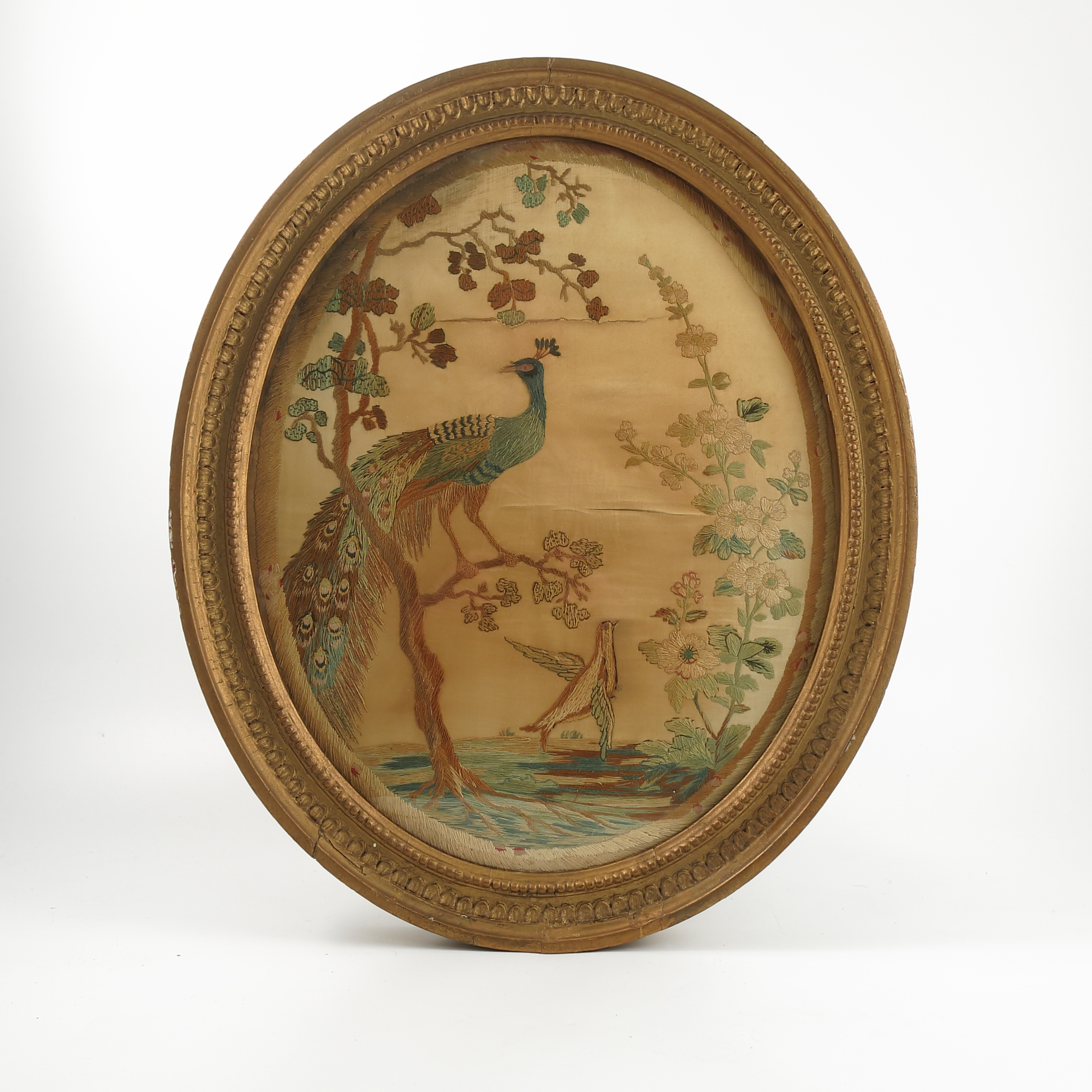 A 19th century oval tapestry picture, on silk, of a peacock in a tree with another bird flying,