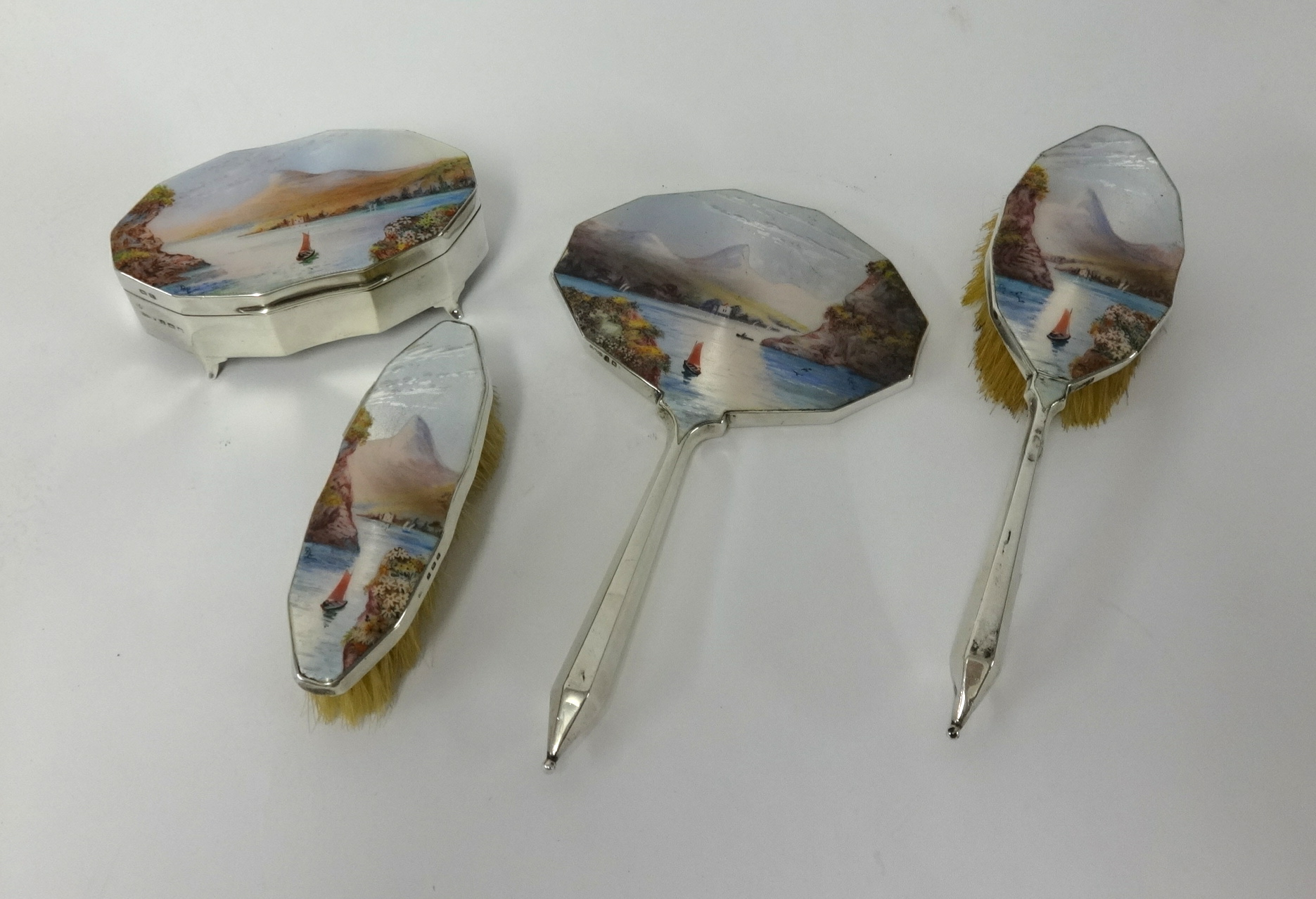 A four piece Geo V silver dressing table set with enamel back depicting a boat, lake and mountain