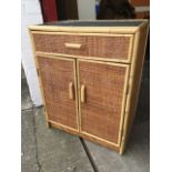 A small cane covered cabinet with panelled tray top above a drawer, with cupboards below.