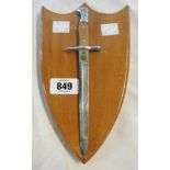 A miniature bayonet, set on a polished wood shield shaped wall plaque