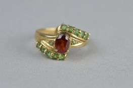 A GARNET AND GREEN STONE 9CT YELLOW GOLD DRESS RING, fancy crossover style, the central oval mixed