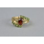 A GARNET AND GREEN STONE 9CT YELLOW GOLD DRESS RING, fancy crossover style, the central oval mixed