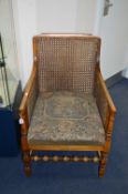 AN EARLY 20TH CENTURY OAK BERGERE ARMCHAIR, with Victorian scene upholstery on turned legs