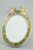 AN OVAL PORCELAIN FRAMED EASEL MIRROR, with cherub mounts and floral encrusted decoration,