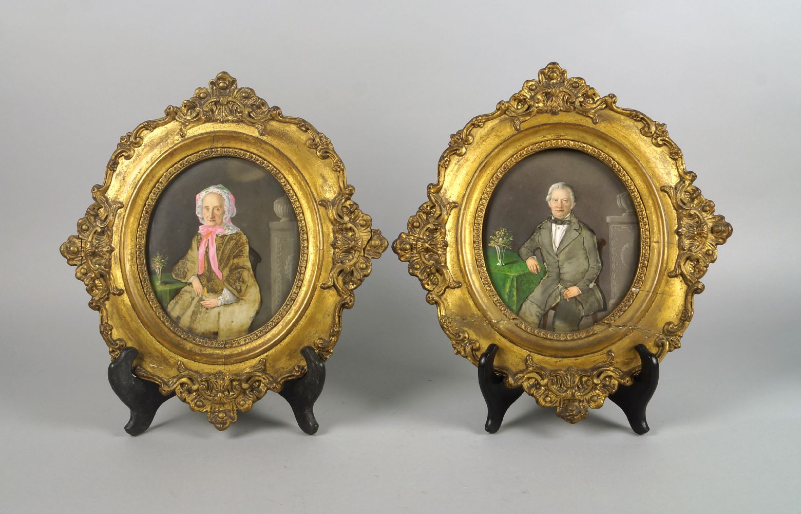 A pair of portraits of a lady and gentleman, 19th century, seated formally at a table with flowers