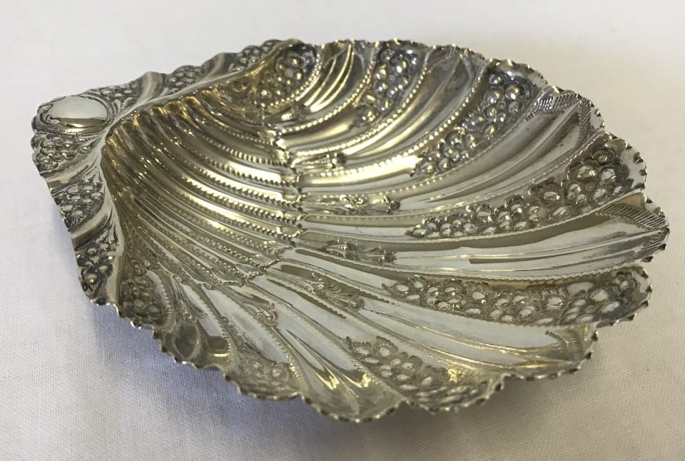 An ornate silver clam shaped dish with floral decoration and 3 conch shaped feet.