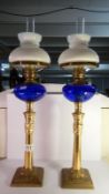 A pair of brass oil lamps with blue glass fonts and white glass shades.