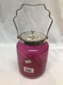 A cranberry glass jar with metal lid and handle.