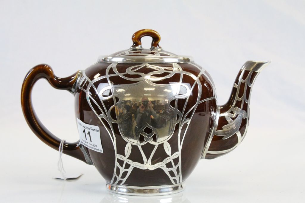 An Art Nouveau treacle glaze teapot with white metal overlay, the central flower motif with