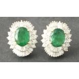 A pair of emerald and diamond ear studs, the oval cut central stones within a band of round and