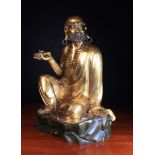 A Gilt Metal Statue of a bearded Arhat (Luohan) sat on a brown patinated rocky outcrop holding a