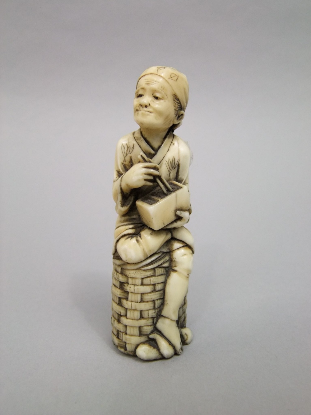 An old Japanese ivory figure of a man, eating with chop sticks while seated on a basket, 10 cm