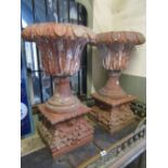 A pair of old weathered terracotta urns on stands with repeating foliate detail raised on square cut