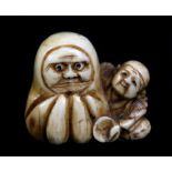 An early 20th century Japanese carved ivory netsuke of a man seated beside a large Daruma doll,