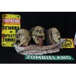 Three rubber zombie busts suspending banner inscribed 'Warning Zombies A Head' attached to rubber