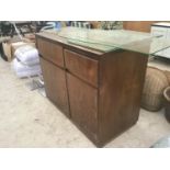 A WOODEN STORAGE CUPBOARD WITH TWO DOORS, A DRAWER AND FLAP AND A QUANTITY OF GLASS