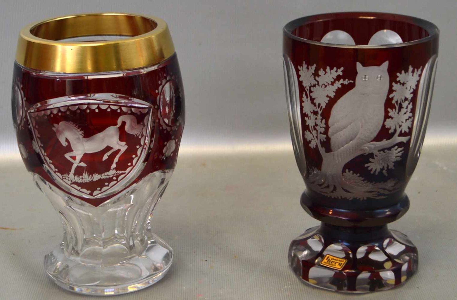 Zwei Becherfarbl. Glas, geschliffen verziert, mit Eule bzw. Reitpferd, mit rotem Überfang, ein