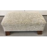 A modern wide foot stool with 4 wooden feet and cream coloured upholstery.