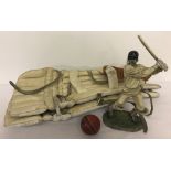A pair of cricket knee pads and red leather cricket ball together with a figurine of a batsman.