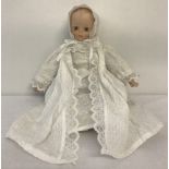A small soft bodied doll with porcelain head, hands and feet. In cotton bonnet and robes.