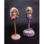 A 1920s composition head on plain wooden stand wearing a knitted cloche hat and a 1930s head on a