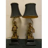 A pair of gilt and metal cherub figured table lamps with black shades on marble bases,