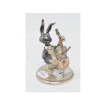 A burnished and silver coloured metal amusing figure, of a hare playing a cello, on a circular base,