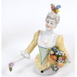 A porcelain half doll, lady wearing a plumed cap and a yellow and green dress, holding a basket of