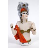 An Art Deco porcelain half doll, Mata Hari, tall grey headdress, red cap and metallic underwear,