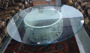 Circular glass topped coffee table with stone carved cylindrical base decorated with figures, 152.
