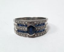 9ct white gold band ring set with a central oval mixed cut sapphire, surmounting three rows of grain