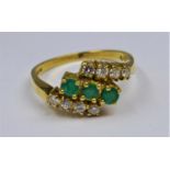 An 18ct yellow gold diamond and emerald ring centrally set with 3 round, faceted emeralds flanked by